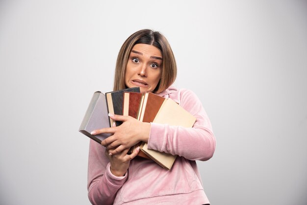 Chica en sudadera rosa sosteniendo y llevando una pesada pila de libros.