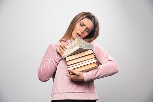 Chica en sudadera rosa sosteniendo una reserva de libros, abriendo uno en la parte superior y leyéndolo.