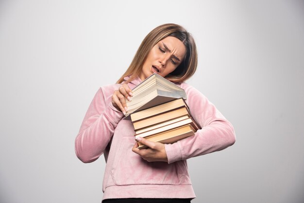 Chica en sudadera rosa sosteniendo una reserva de libros, abriendo uno en la parte superior y leyéndolo.