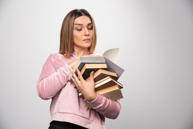 Chica en sudadera rosa sosteniendo una reserva de libros, abriendo uno en la parte superior y leyéndolo.