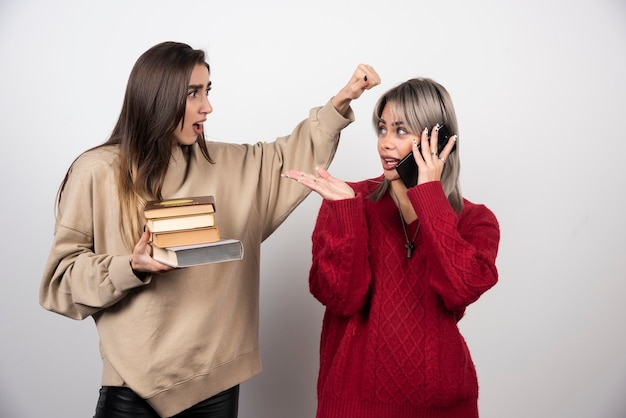 Chica en suéter beige sosteniendo libros mientras otra chica habla por teléfono.