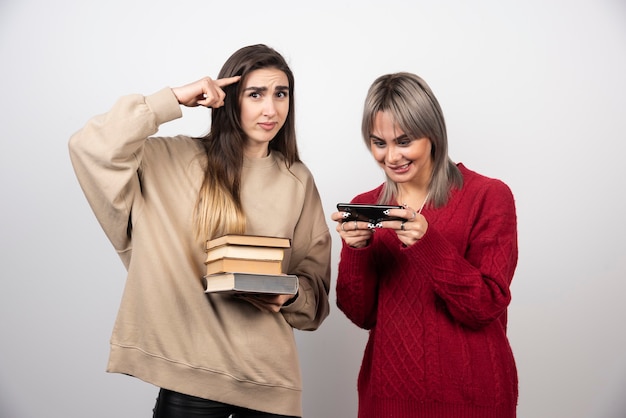 Chica en suéter beige sosteniendo libros mientras otra chica mirando el teléfono.