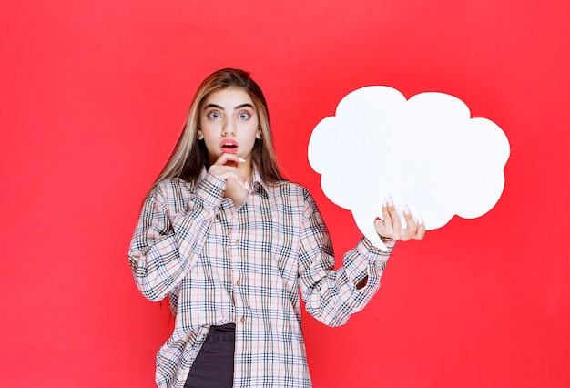 Chica en suéter cálido sosteniendo un tablero de ideas con forma de nube y parece sorprendida