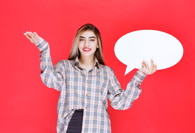 Chica en suéter de cuadros sosteniendo un tablero de ideas ovale y apuntando a los participantes