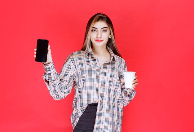 Chica en suéter de cuadros sosteniendo una taza de bebida y mostrando su teléfono inteligente