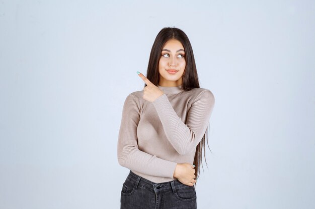 Chica en suéter gris mostrando algo a la izquierda.