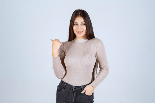 Chica en suéter gris mostrando algo a la izquierda.