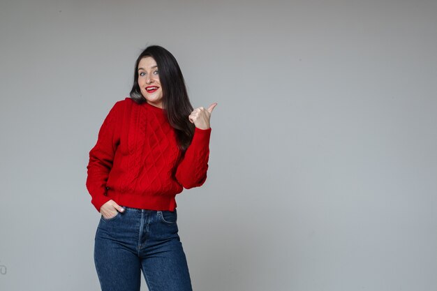 Chica en suéter rojo y jeans apuntando al espacio en blanco.