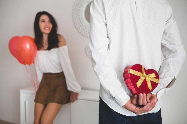 Chica sujetando globos con forma de corazón mientras su novio tiene un regalo para ella a la espalda