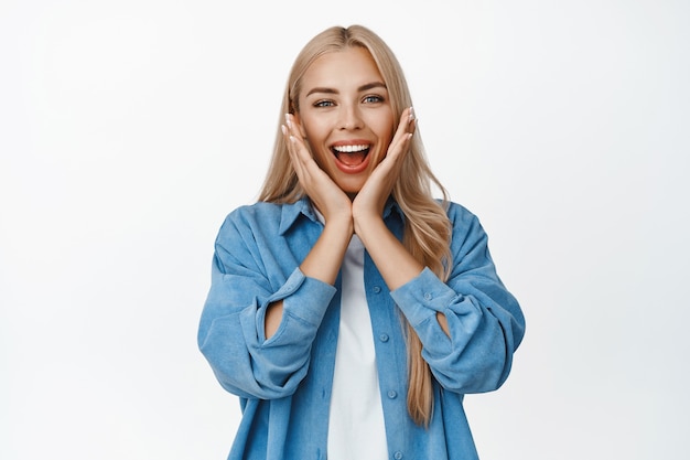 Chica súper feliz regocijándose, tomados de la mano cerca de la cara y sonriendo sorprendido, recibe grandes buenas noticias, de pie en blanco.