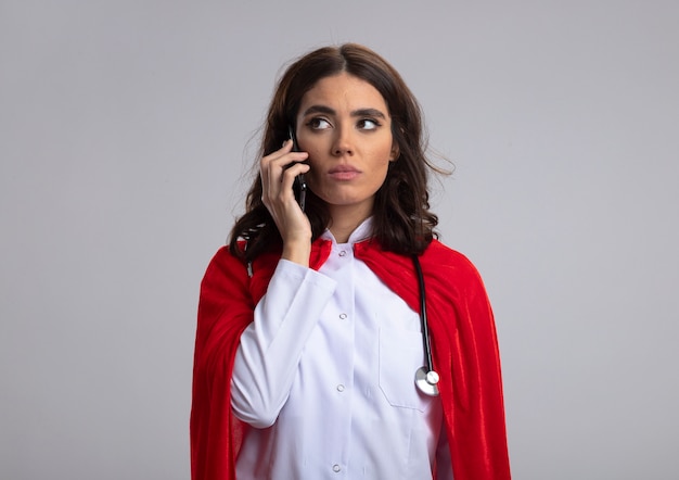 Chica de superhéroe caucásica confiada en uniforme médico con capa roja y estetoscopio habla por teléfono mirando al lado aislado en la pared blanca con espacio de copia