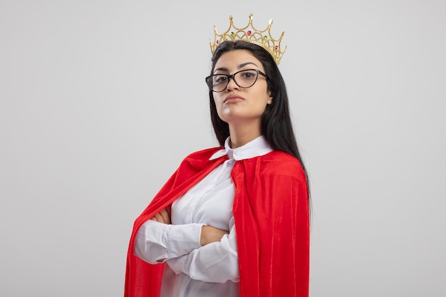 Foto gratuita chica de superhéroe caucásica joven confiada con gafas y corona de pie con postura cerrada en vista de perfil mirando a cámara aislada sobre fondo blanco con espacio de copia