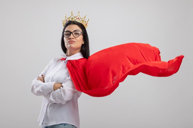 Foto gratuita chica de superhéroe caucásica joven confiada con gafas y corona de pie con postura cerrada en vista de perfil mirando a cámara aislada sobre fondo blanco