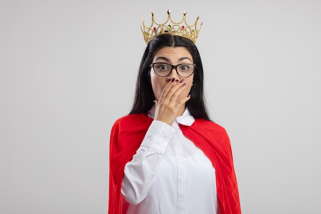 Chica superhéroe caucásica joven sorprendida con gafas y corona mirando a cámara manteniendo la mano en la boca aislada sobre fondo blanco con espacio de copia