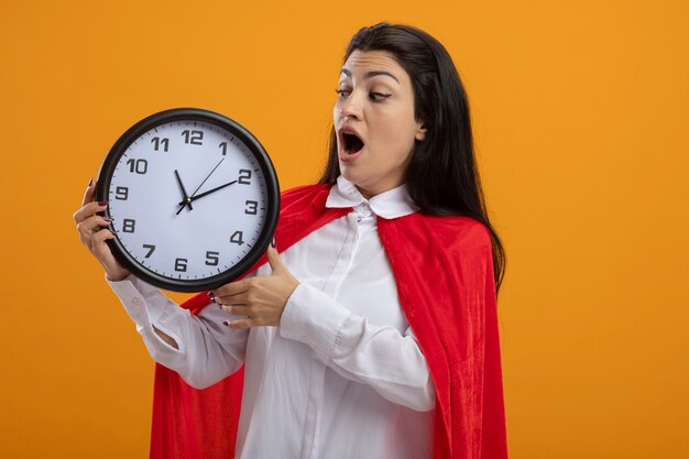 Chica de superhéroe caucásica joven sorprendida sosteniendo y mirando el reloj aislado en la pared naranja con espacio de copia