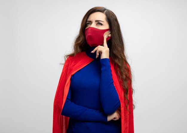 Chica de superhéroe caucásica pensativa con capa roja con máscara protectora roja pone la mano en la barbilla mirando al lado aislado en la pared blanca con espacio de copia