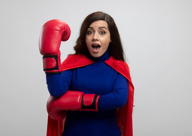 Chica de superhéroe caucásica sorprendida con capa roja con guantes de boxeo tiene la mano en blanco