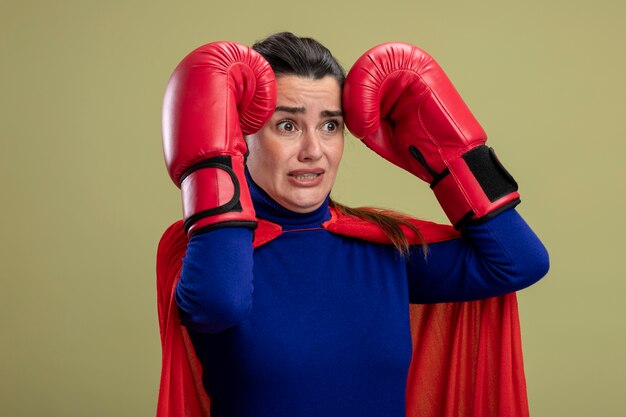 Chica superhéroe joven asustada mirando al lado con guantes de boxeo poniendo las manos en la cabeza aislada en verde oliva