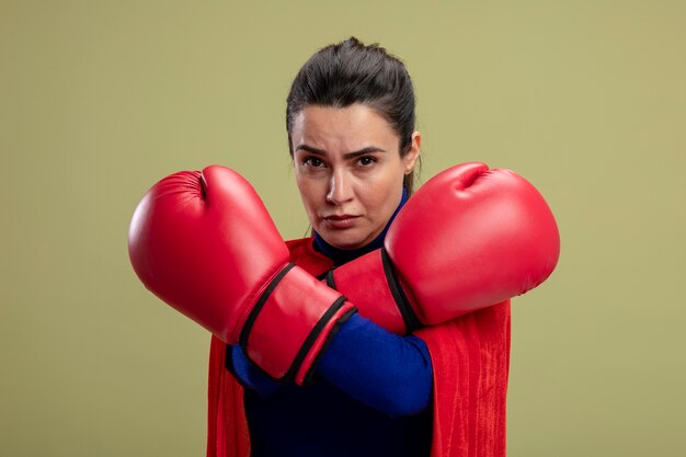 Chica de superhéroe joven confiada con guantes de boxeo cruzando las manos aisladas sobre fondo verde oliva