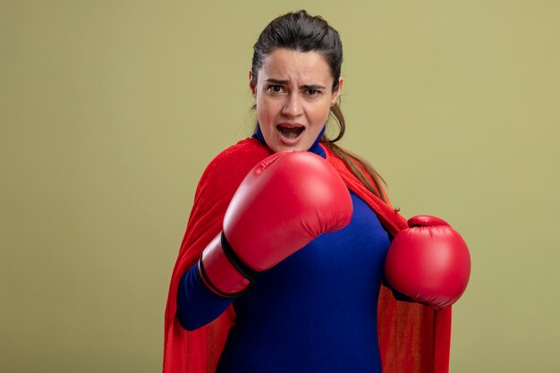 Chica de superhéroe joven confiada con guantes de boxeo de pie en pose de combate aislado sobre fondo verde oliva