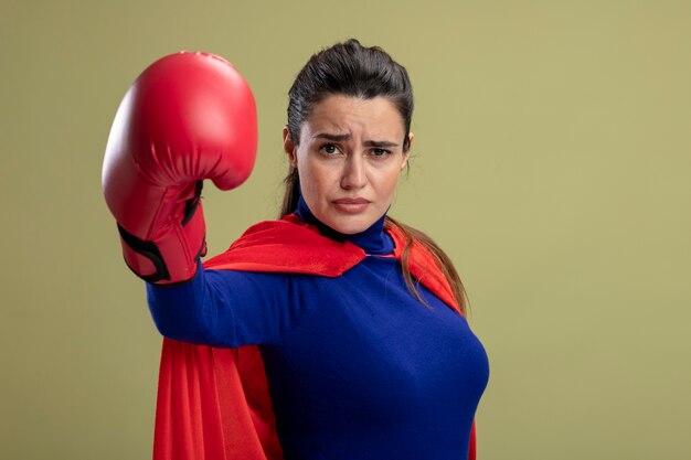 Chica de superhéroe joven confiada con guantes de boxeo sosteniendo la mano a la cámara aislada sobre fondo verde oliva