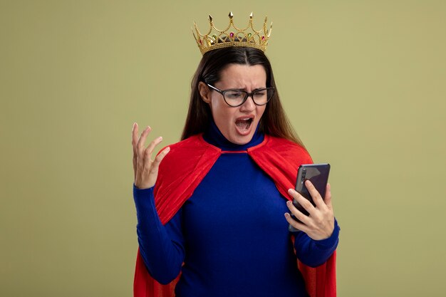 Chica superhéroe joven enojada con gafas y corona sosteniendo y mirando el teléfono aislado sobre fondo verde oliva