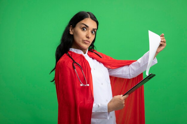 Chica superhéroe joven sorprendida mirando al frente vistiendo bata médica con estetoscopio sosteniendo y volteando a través del portapapeles aislado en verde