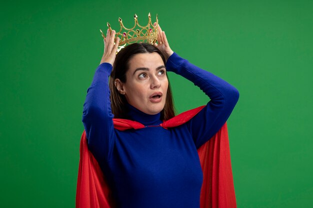 Chica superhéroe joven sorprendida mirando al lado con corona poniendo corona en la cabeza aislada en verde