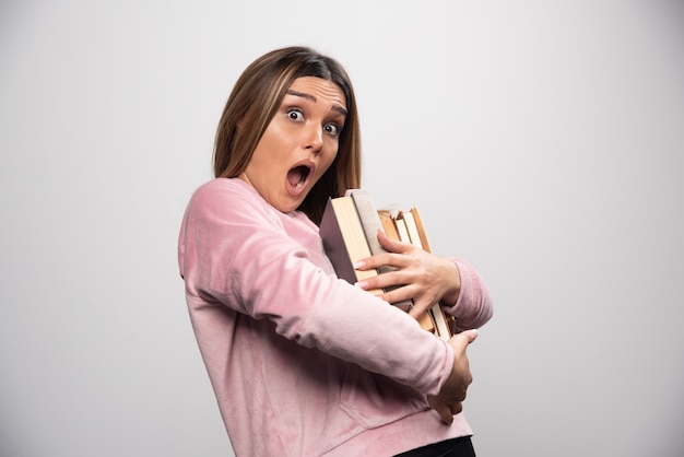 Chica en swaetshirt rosa sosteniendo y llevando una pesada pila de libros.