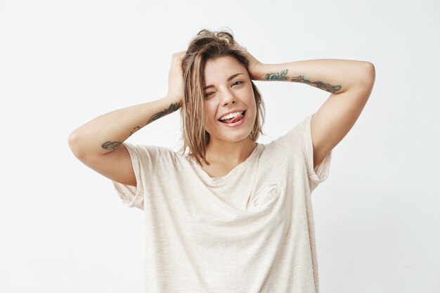 Chica tatuada fresca alegre sonriendo guiñando un ojo mostrando la lengua tocando el cabello.