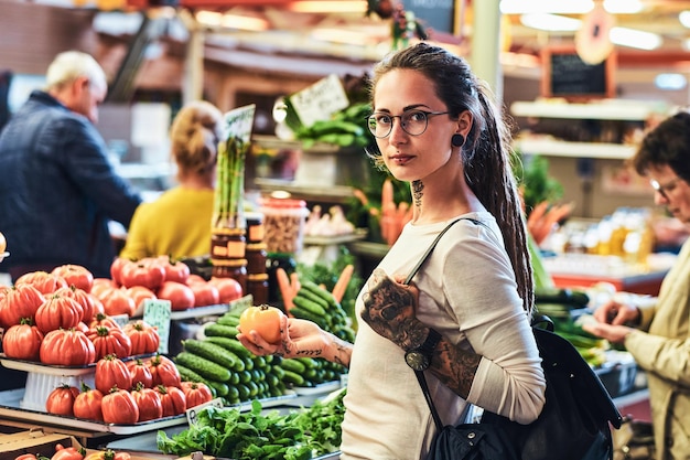 Chica tatuada pensativa está eligiendo cuidadosamente tomates en el mercado de un agricultor local.