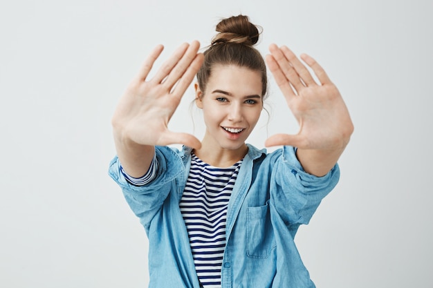 Foto gratuita chica te vigila. retrato de mujer caucásica alegre emotiva tirando de las manos hacia la cámara, haciendo triángulo y mirando a través de él con una amplia sonrisa y mirada de complicidad, de pie sobre la pared gris