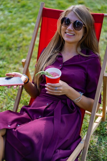 chica con un teléfono inteligente y un cóctel en la naturaleza en una tumbona. Día libre de verano.