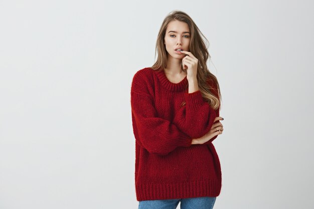 Chica en la tienda intenta elegir zapatos nuevos. Foto de estudio de una chica urbana guapa con un suéter rojo suelto de pie con el brazo cruzado y la mano en el labio, eligiendo entre las variantes mientras se mira