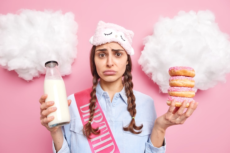 Foto gratuito chica tiene coletas se ve con expresión de mal humor quiere comer deliciosas donas al horno come postre dulce y bebe leche fresca viste ropa casual aislado en rosa