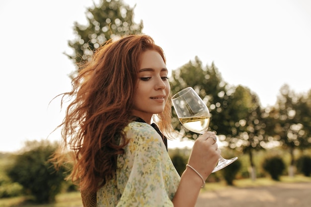 Chica tierna con cabello largo jengibre y pecas encantadoras en ropa verde de verano de moda mirando hacia abajo y sosteniendo un vaso con vino al aire libre