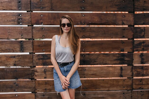 Chica tímida en falda de mezclilla de moda posando junto a la pared de madera. Retrato al aire libre de adorable dama caucásica con peinado recto.