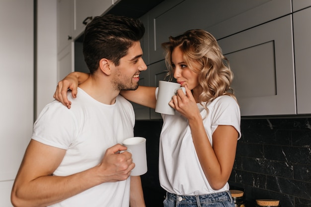 Chica tocando juguetonamente a marido y tomando café. Retrato interior de una pareja romántica disfrutando del desayuno juntos.