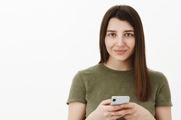 Chica tomando notas en el teléfono inteligente que parece lista y positiva con tierna sonrisa linda mientras espera el comando sonriendo de pie despreocupado y tímido contra la pared gris con el teléfono móvil en las manos