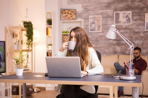 Chica tomando un sorbo de café mientras trabaja en la computadora portátil en la oficina en casa. Novio relajándose en el sofá en el fondo.