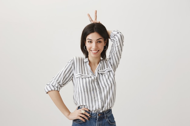 Foto gratuita chica tonta alegre mostrando gesto de orejas de conejo detrás de la cabeza y sonriendo