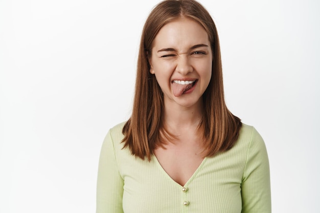 Chica tonta y feliz, muestra la lengua y guiña un ojo a la cámara, sonriendo despreocupada, disfrutando de los días positivos de verano, de pie en una blusa verde contra un fondo blanco. copia espacio