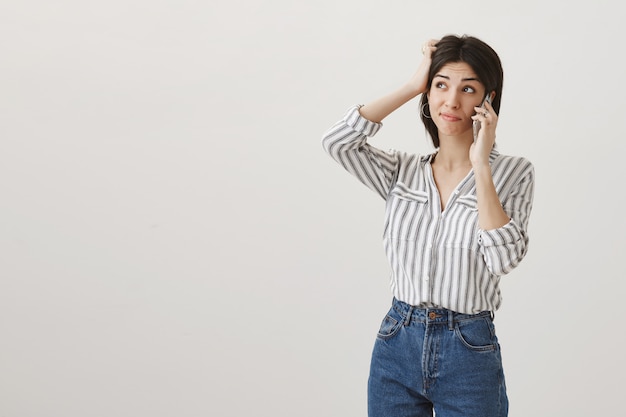 Chica tonta indecisa rascándose la cabeza insegura, hablando por teléfono, tomando decisiones y mirando en la esquina superior izquierda