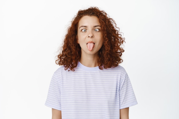 Foto gratuita chica tonta y rizada con el pelo rojo mostrando la lengua, caras divertidas, de pie en blanco con camiseta casual.