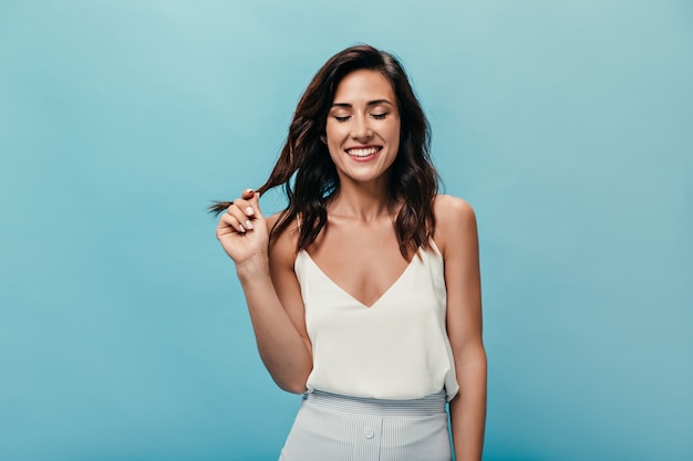 Chica en top blanco coquetamente toca su cabello y sonríe sobre fondo aislado