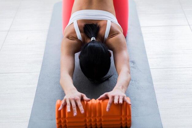Una chica con un top blanco y mallas de coral usa un rodillo de espuma para relajarse después de una sesión de fitness.