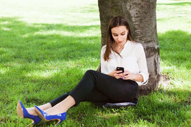 Chica trabaja con su teléfono sentado bajo el árbol