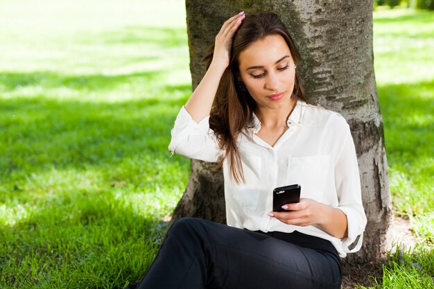 Chica trabaja con su teléfono sentado bajo el árbol