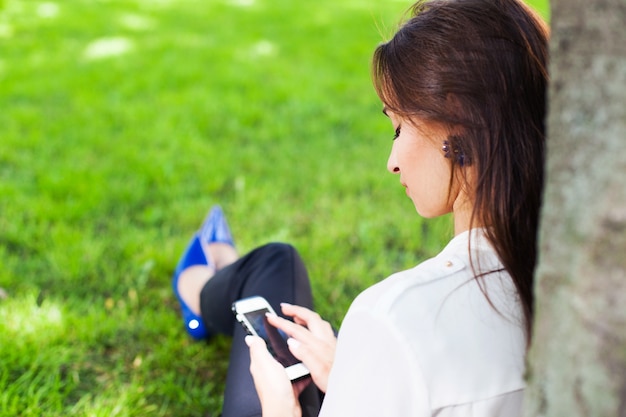 Chica trabaja con su teléfono sentado bajo el árbol