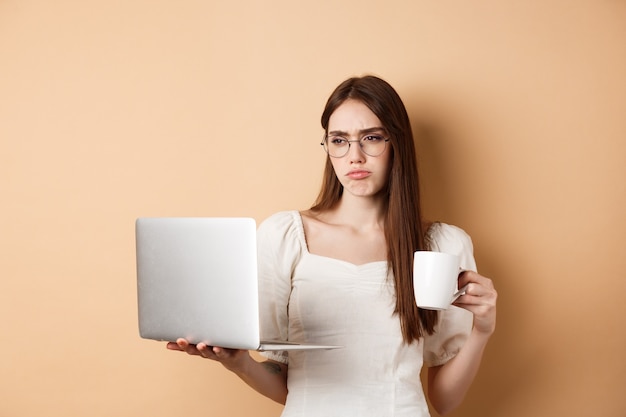 Chica trabajadora cansada mirando la computadora portátil aburrida bebiendo café mientras usa la computadora para trabajar de pie en ...